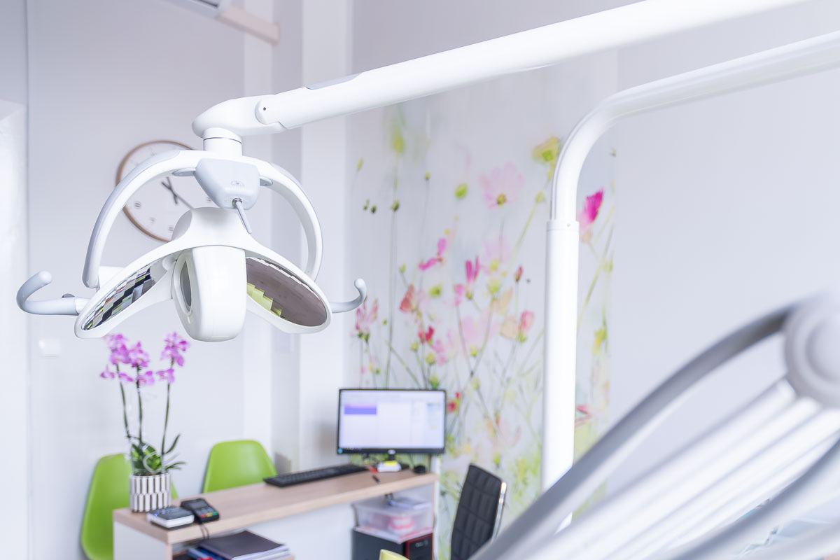 Jasny gabinet stomatologiczny z lampą zabiegową, unitem dentystycznym i stanowiskiem komputerowym.